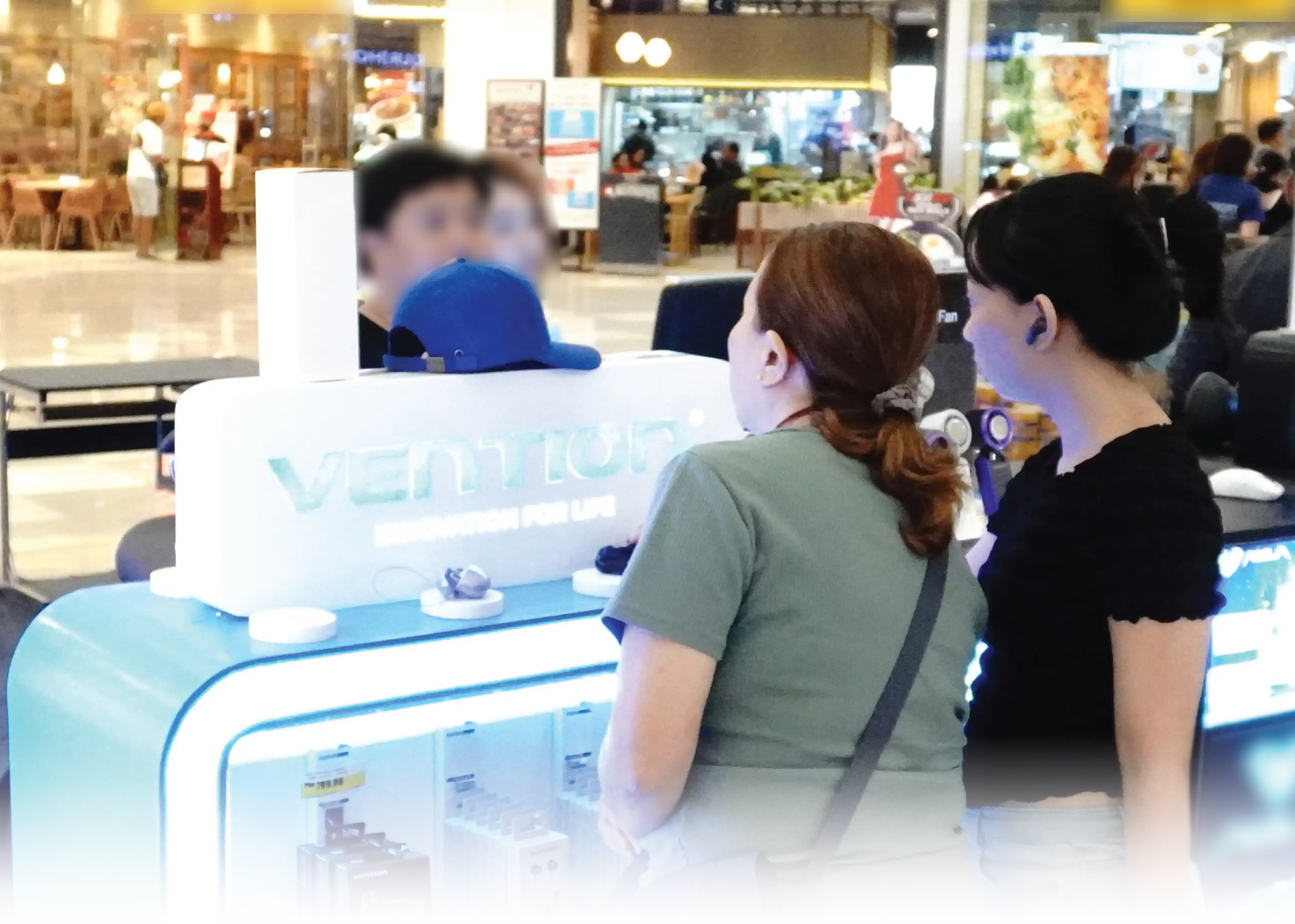Customers browsing a brightly lit blue and white Vention retail display kiosk inside a shopping mall near an Office Warehouse.