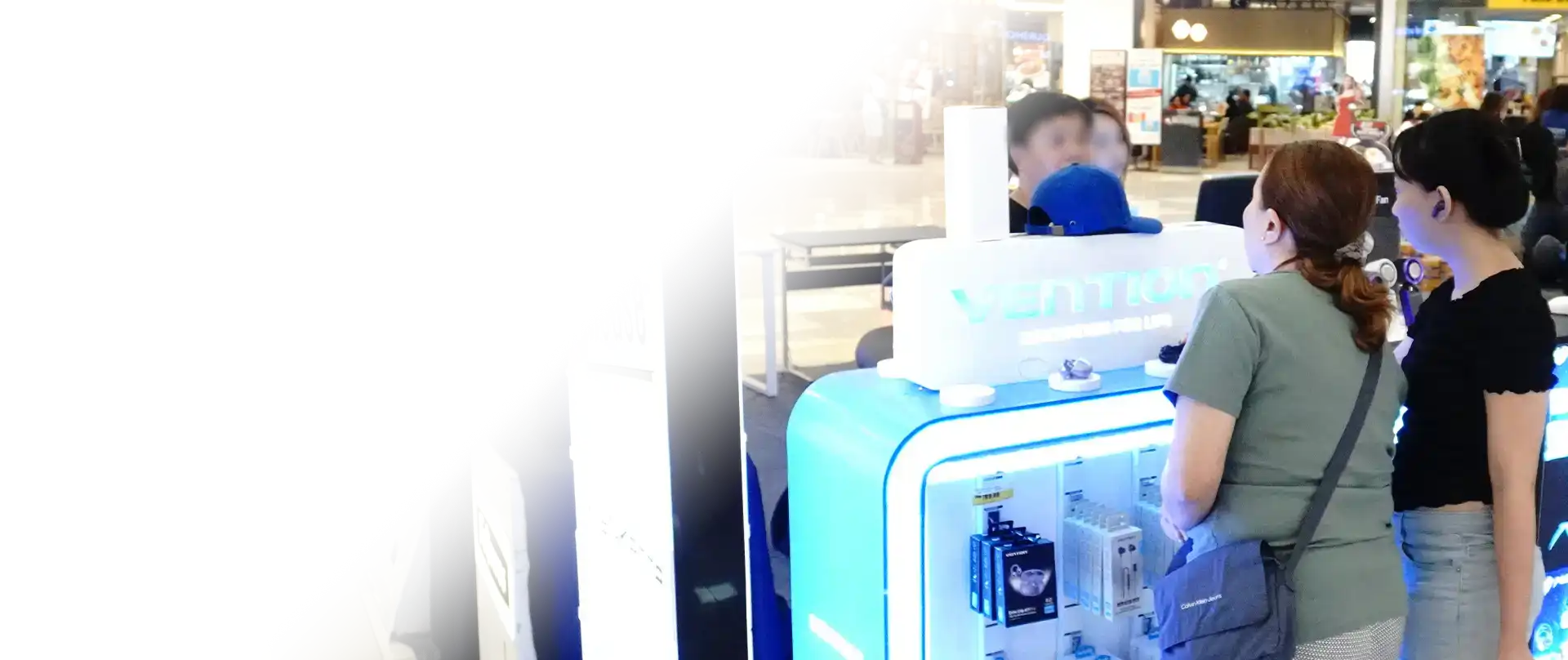 Customers browsing a brightly lit blue and white Vention retail display kiosk inside a shopping mall near an Office Warehouse.
