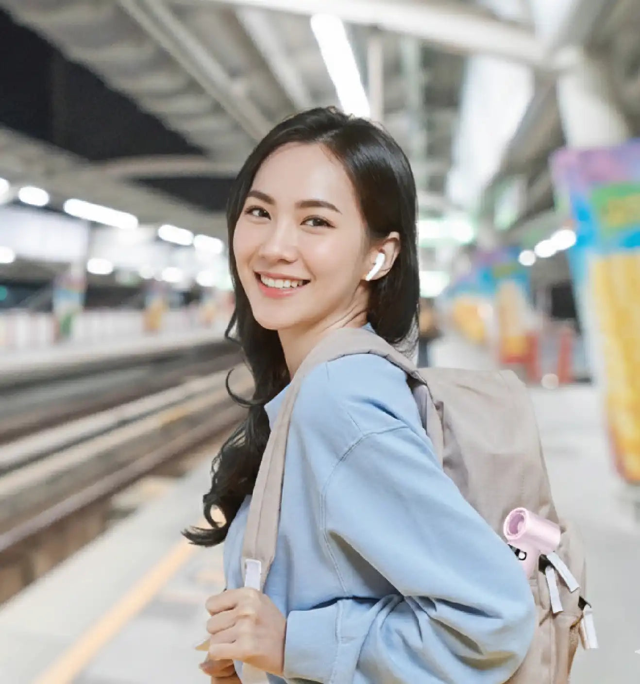 A smiling young woman wearing white wireless earbuds and a backpack stands on a train station platform, representing a modern lifestyle and travel convenience.